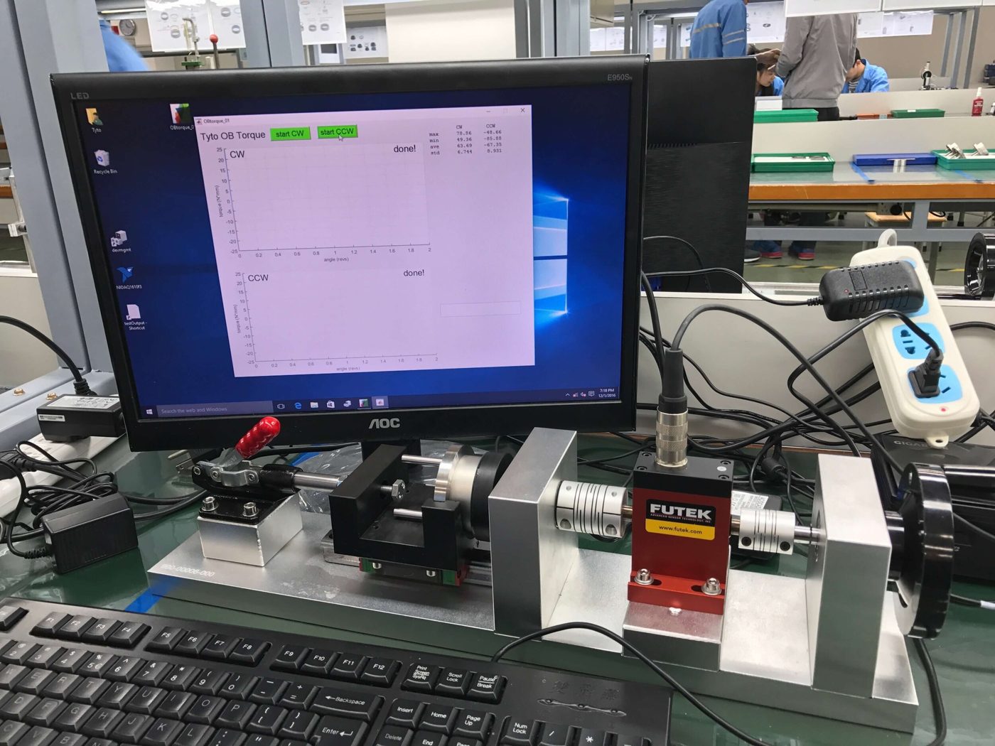Bezel torque fixture: a red FUTEK torque sensor inline on a rail with a toggle clamp and crank, a PC showing the torque program behind it