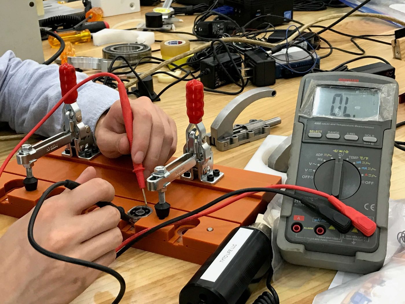 Orange fixture board with toggle clamps holding bolt backs while an operator probes the pogo pins with a multimeter
